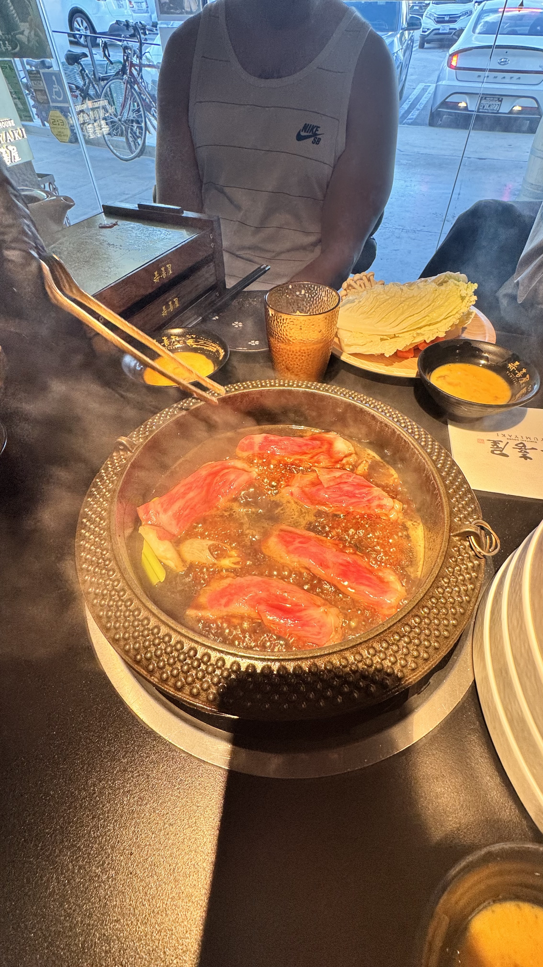 A hot pot meal with thinly sliced meat cooking in broth, surrounded by various side dishes and drinks on a table