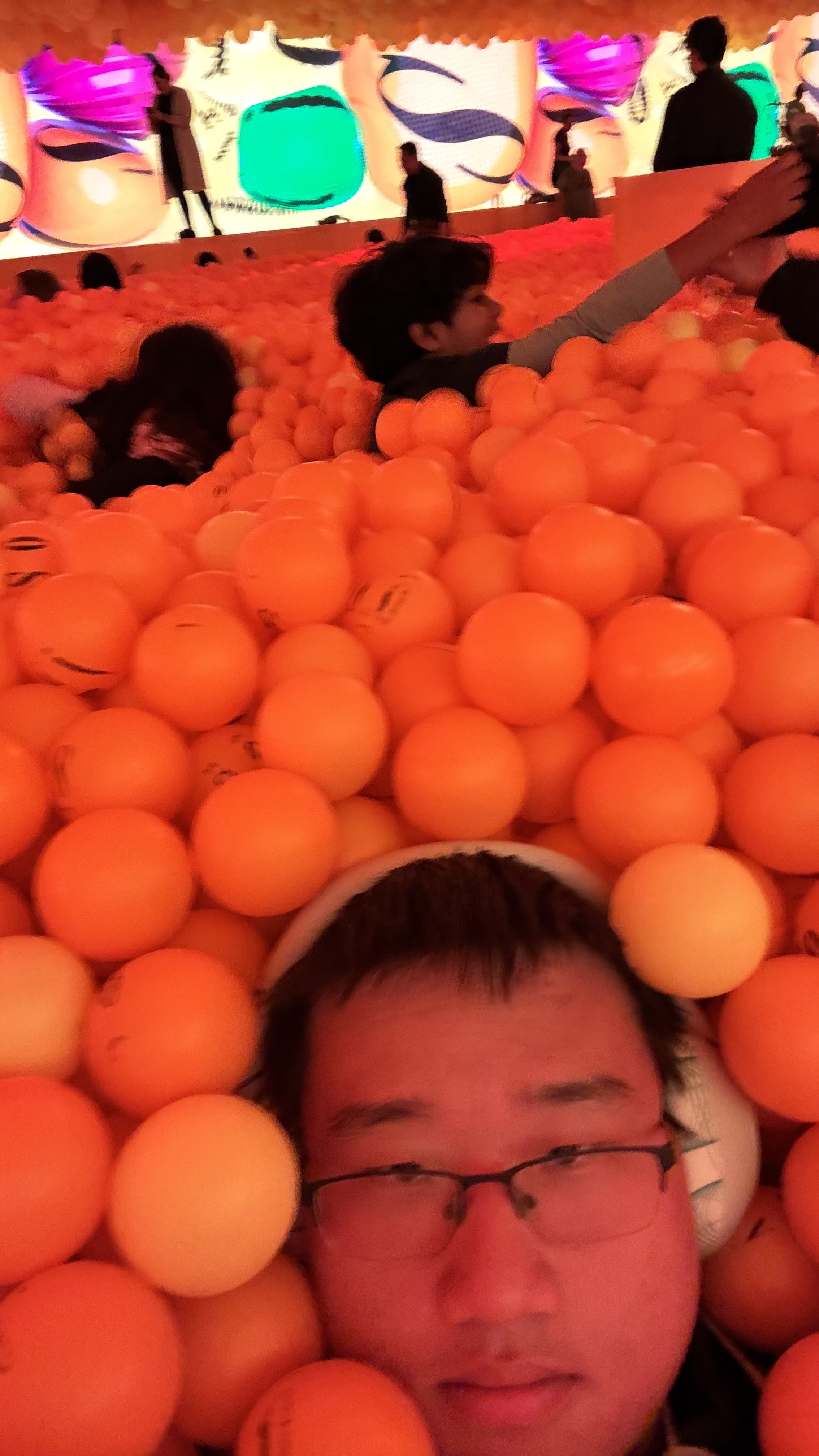 A person is partially submerged in a large pit filled with orange balls, with others visible in the background