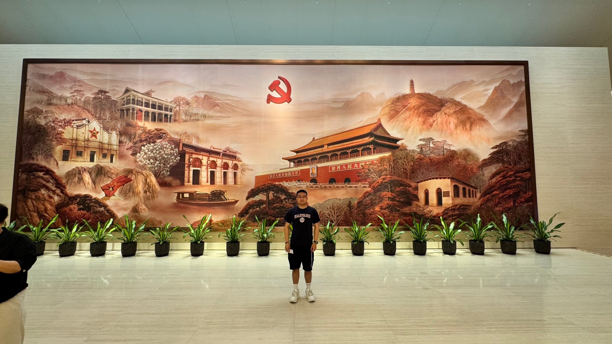 A person stands in front of a large mural depicting traditional Chinese architecture and landscapes, with a row of potted plants below