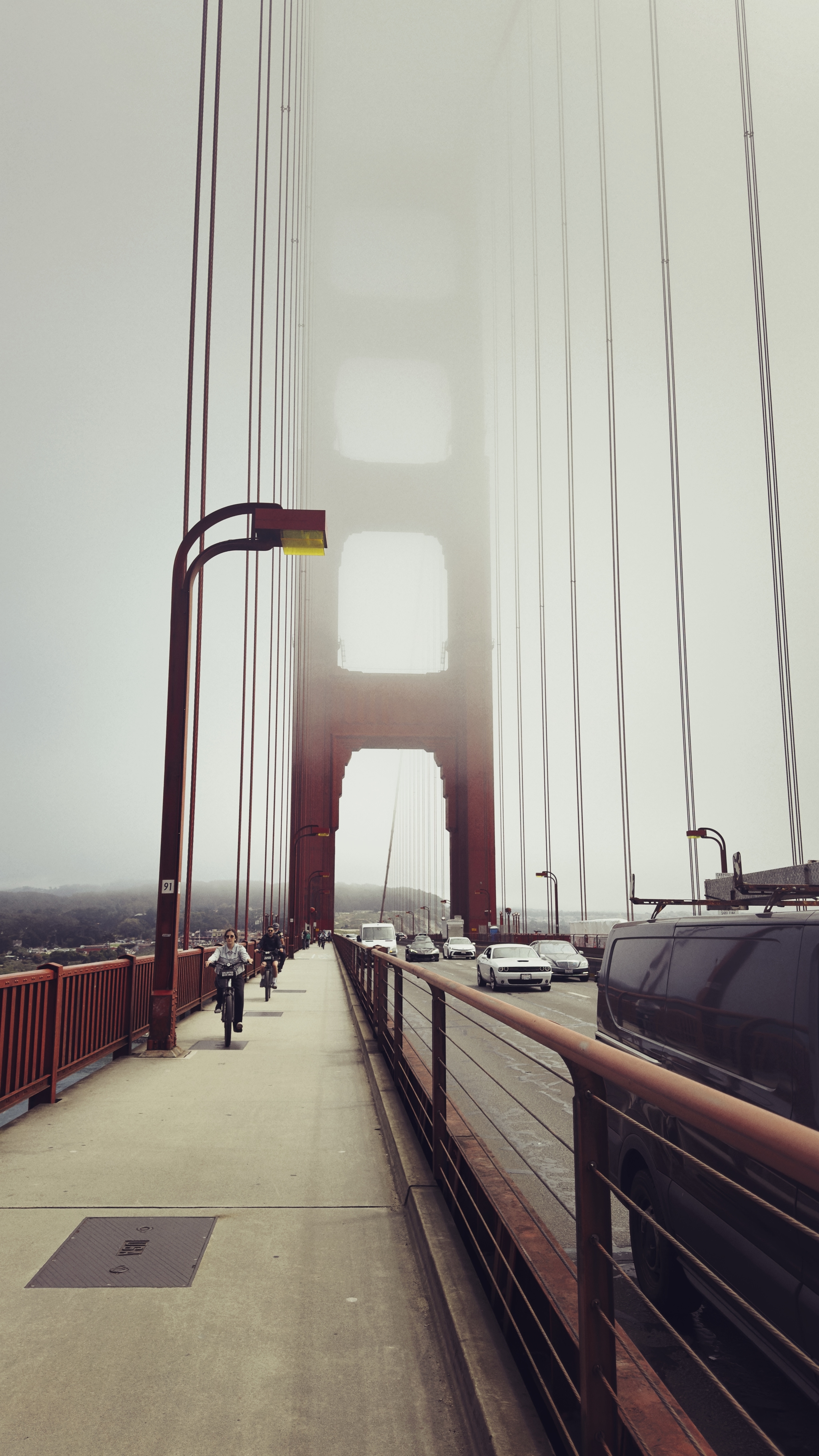 A foggy view of a suspension bridge with pedestrians walking on the sidewalk and vehicles on the road