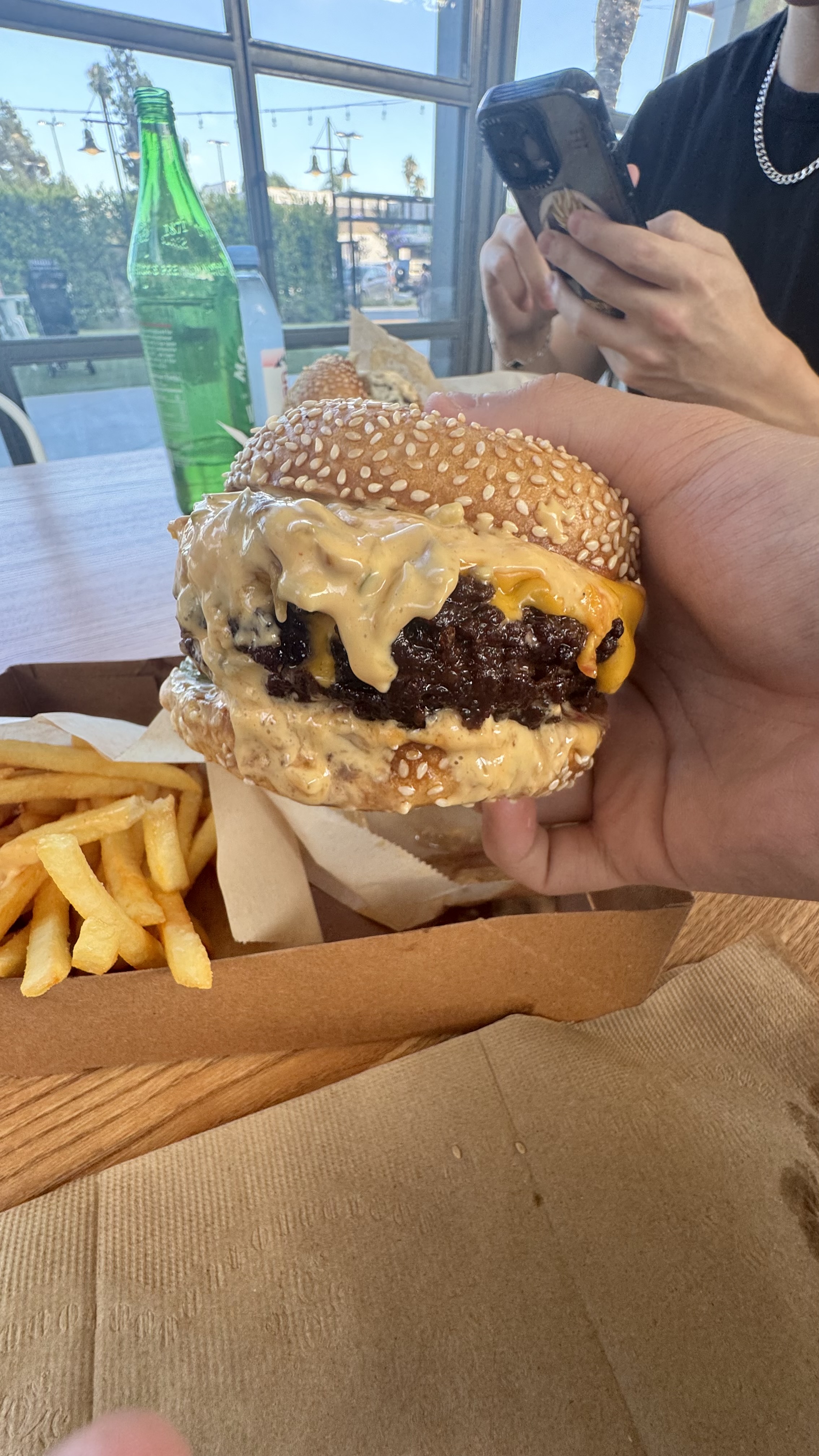 A person holding a cheeseburger with a sesame seed bun, accompanied by a serving of fries on a tray. Another person is using a smartphone in the background, and a green bottle is on the table
