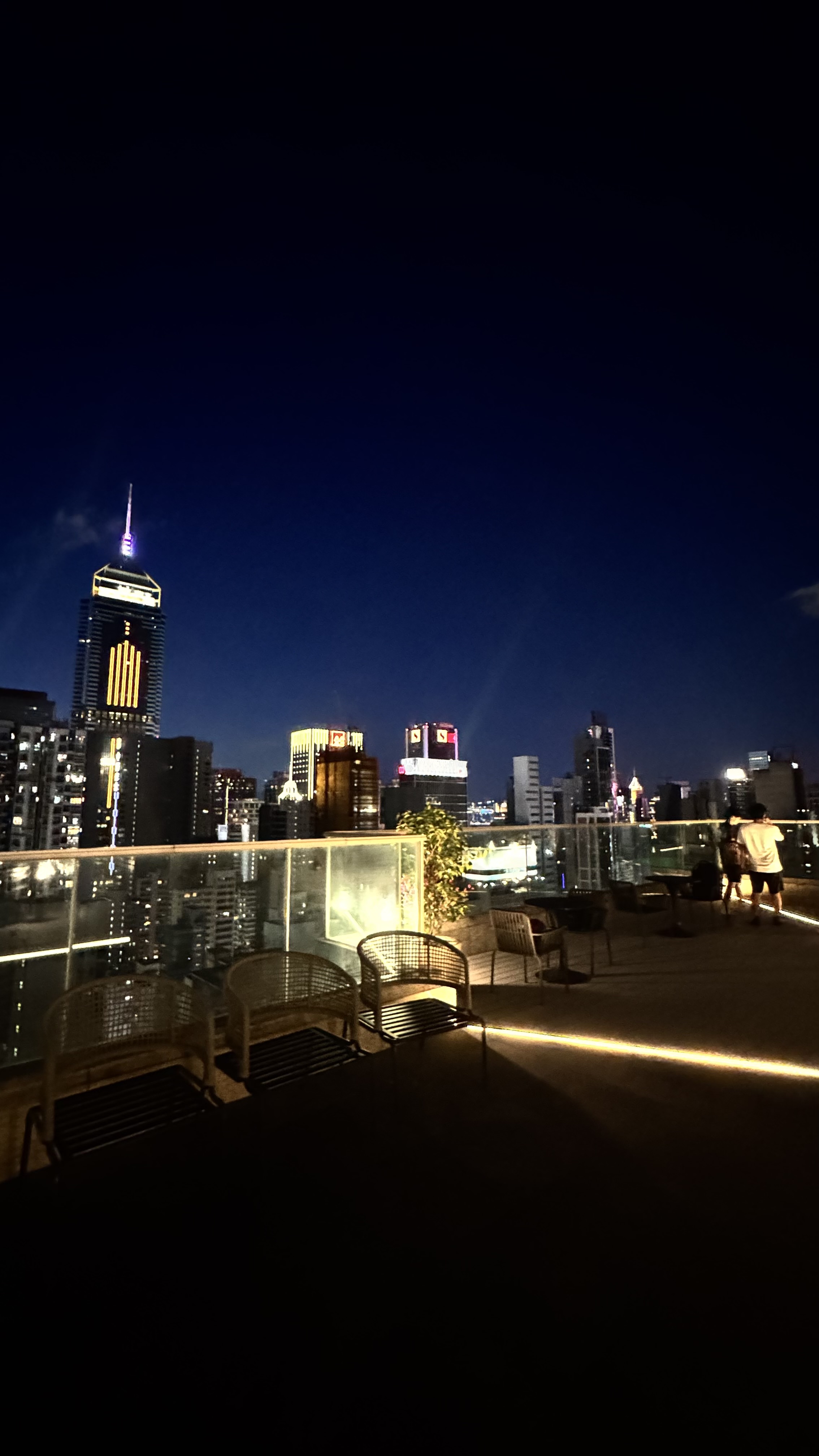 A city skyline at night with illuminated skyscrapers and a rooftop terrace featuring seating and ambient lighting