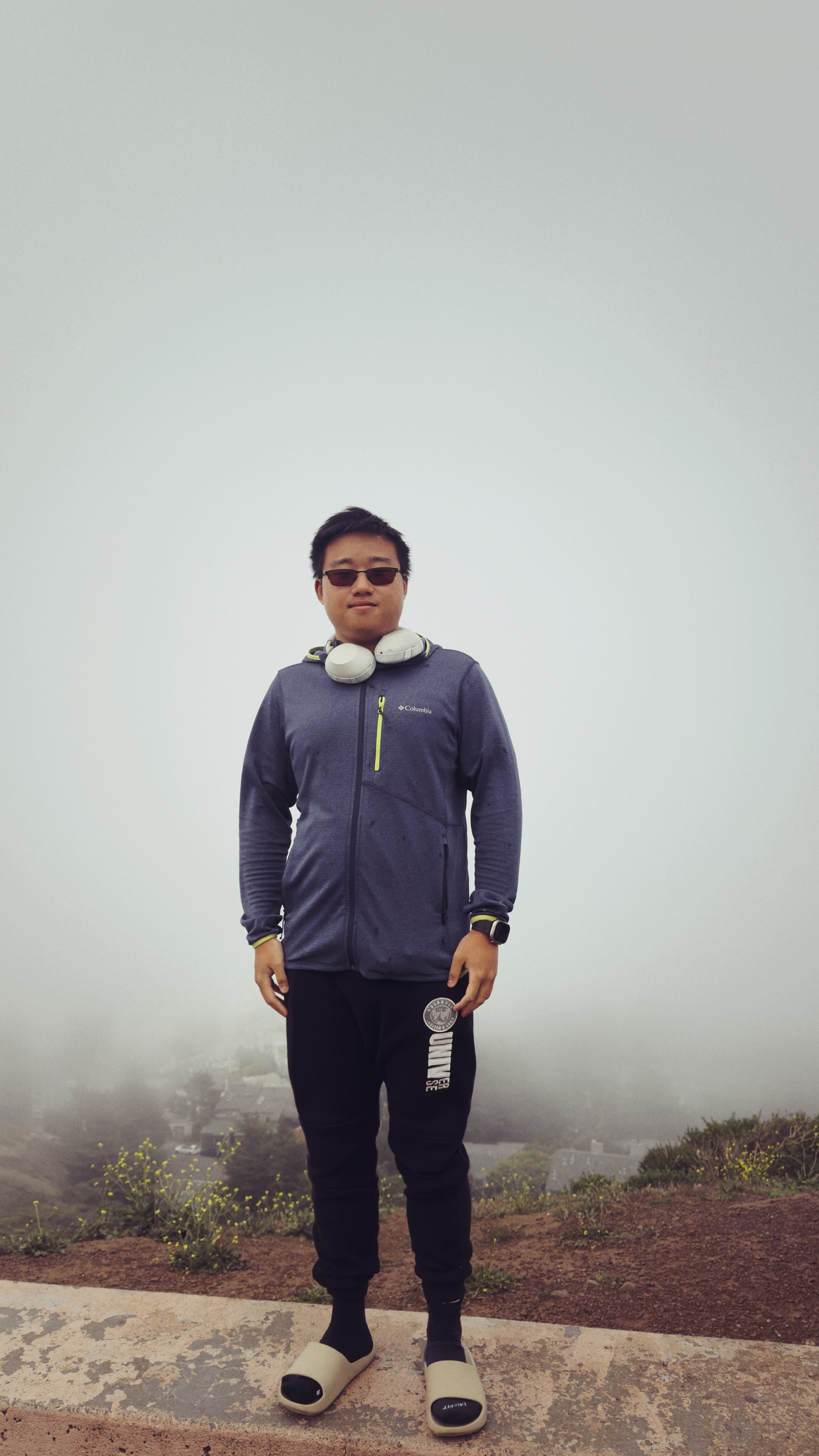 A person wearing a jacket, headphones, and sunglasses stands on a foggy outdoor path, holding a water bottle