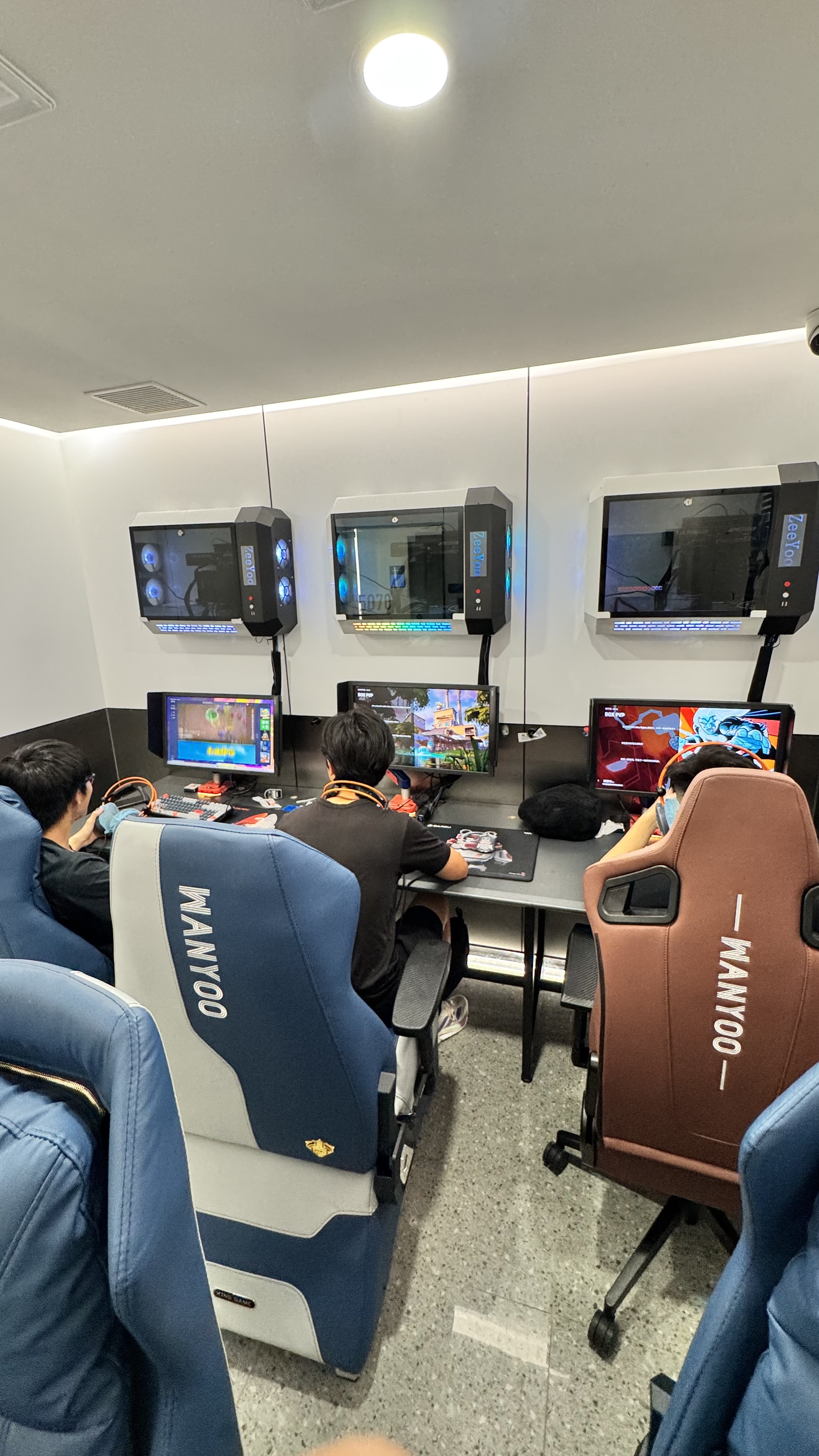 A gaming room with three people seated in ergonomic chairs, playing video games on multiple monitors mounted on the wall