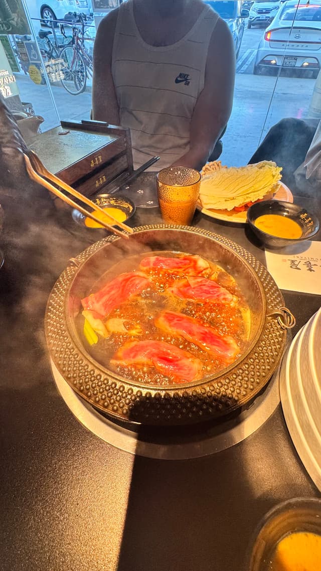 A hot pot meal with thinly sliced meat cooking in broth, surrounded by various side dishes and drinks on a table