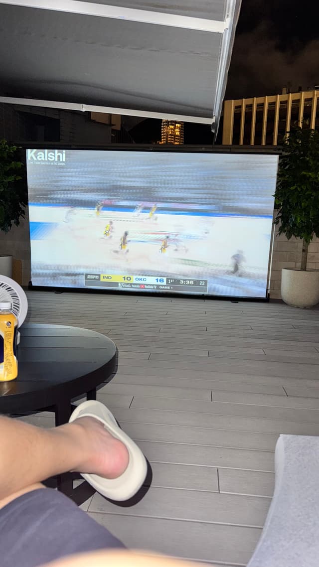 A person relaxing on a patio with feet up, watching a sports game projected on a screen