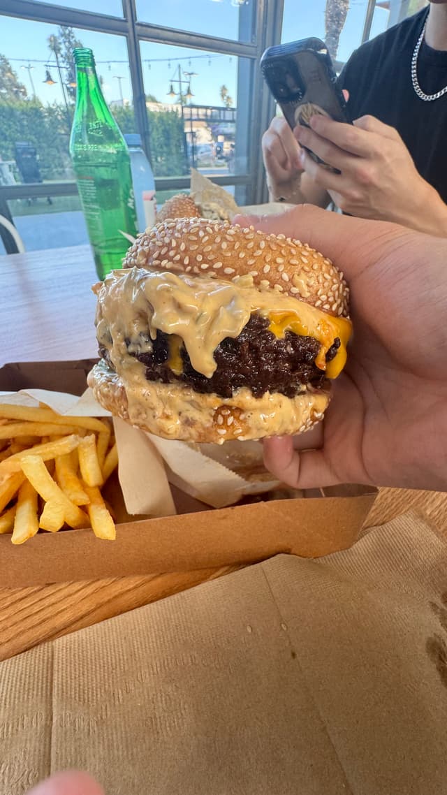 A person holding a cheeseburger with a sesame seed bun, accompanied by a serving of fries on a tray. Another person is using a smartphone in the background, and a green bottle is on the table
