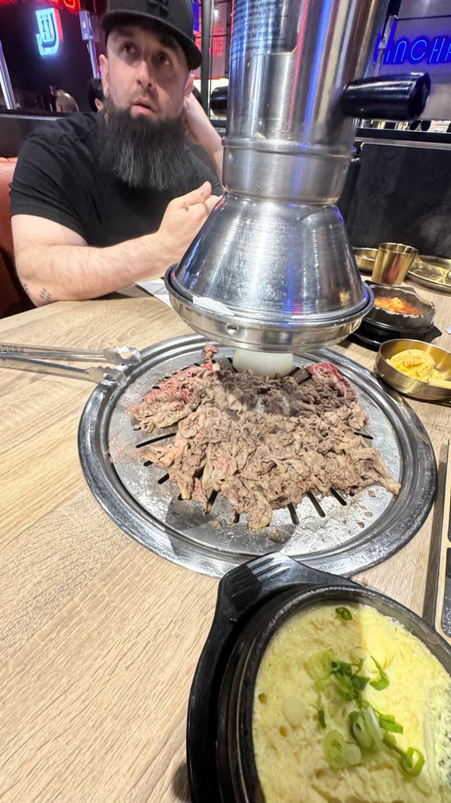 A man sitting at a table in a restaurant with a grill in the center, cooking sliced meat. A metal exhaust hood is positioned above the grill, and there are side dishes and a bowl of soup on the table