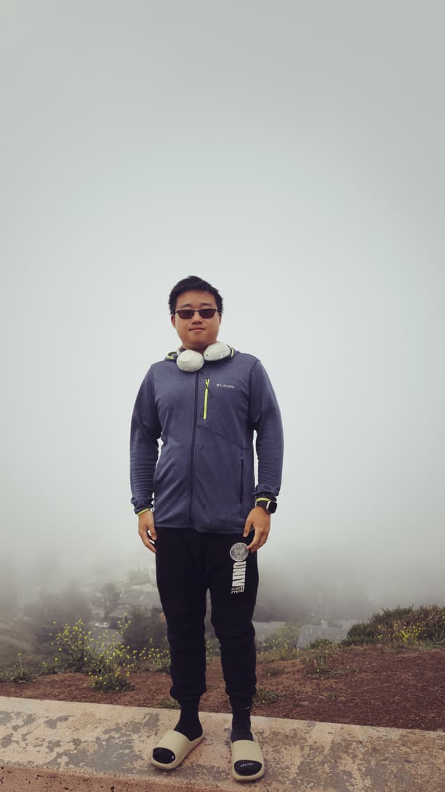 A person wearing a jacket, headphones, and sunglasses stands on a foggy outdoor path, holding a water bottle