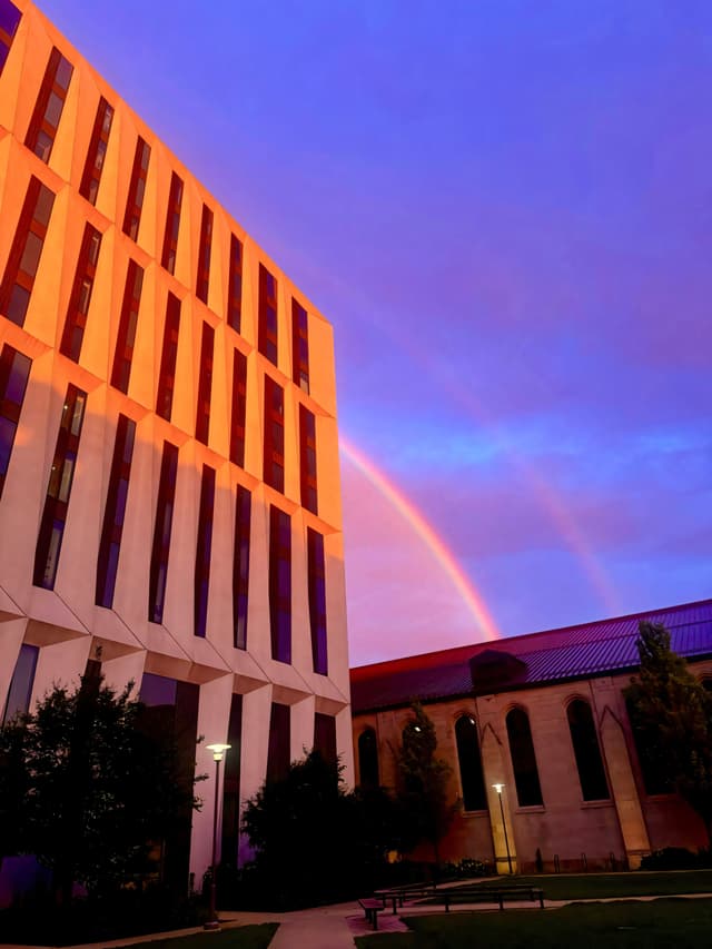 A modern building with vertical windows is illuminated by a warm light, set against a vibrant sky featuring a double rainbow
