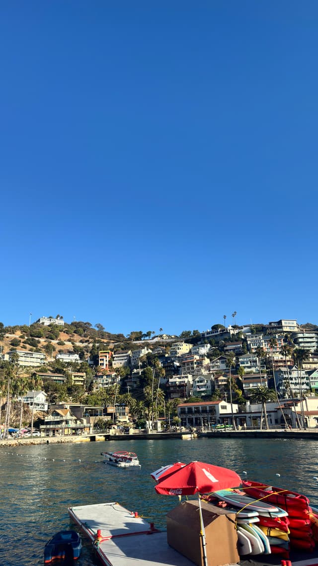 A coastal town with hillside houses, a calm body of water, and a dock with a red umbrella and kayaks